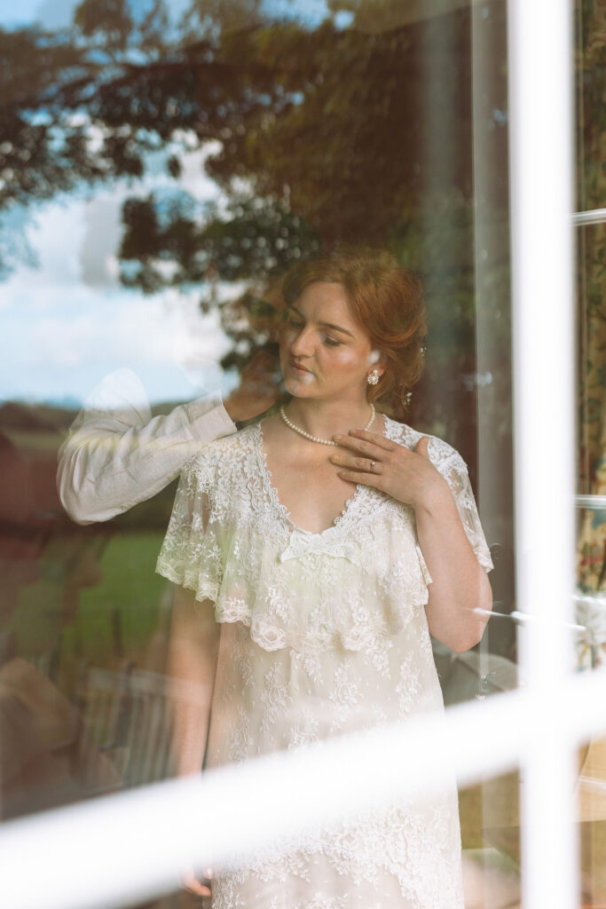 couple gets ready together on their elopement day at teasses estate in scotland, you can see them through the window of their cottage with the trees reflecting on the window and the bride holding pearls as the groom clasps them on her neck
