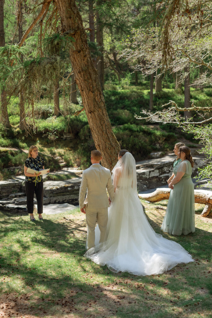 elopement day at Linn of Quoich next to the river where the bride and groom hold hands and two sisters stand nearby as witnesses 