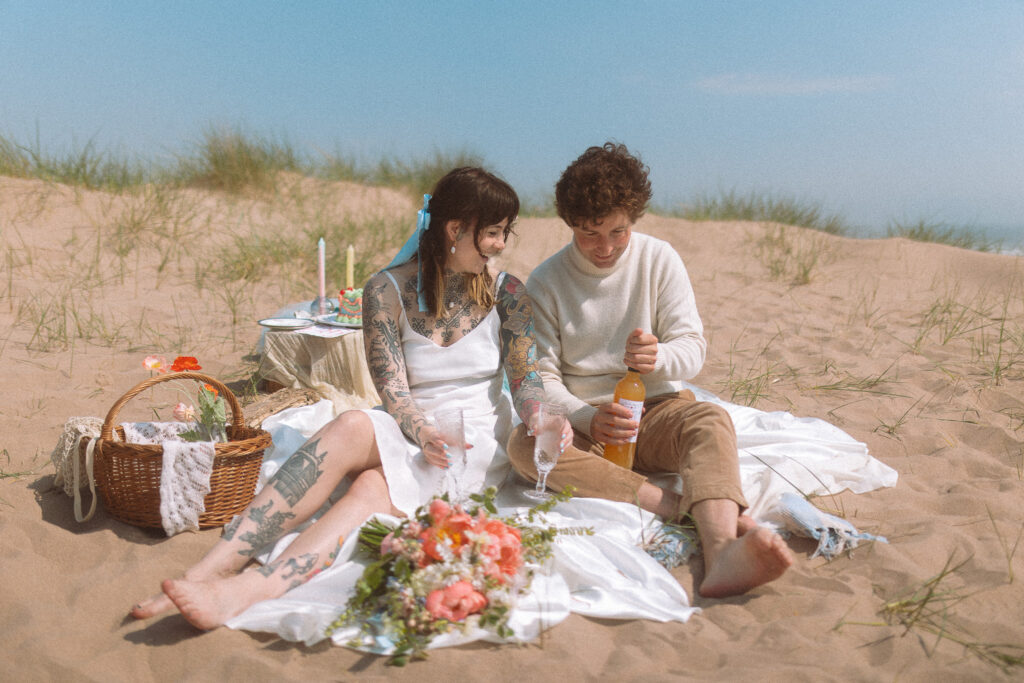 bride and groom pop some champagne after they say their vows on a beach wedding. there are pastel colour flowers in her bouquet and a picnic basket next to them